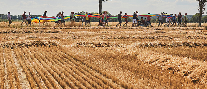 Teilnehmer des CSD Wendland laufen mit Regenbogenfahne neben Kornfeld